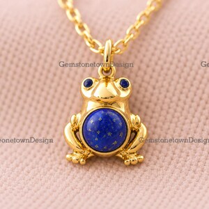 Può includere: Un ciondolo dorato a forma di rana appeso a una catenina abbinata. La rana presenta una grande pietra rotonda di lapislazzuli blu intenso sulla pancia e due pietre blu più piccole per gli occhi. Il testo GemstonetownDesign è visibile.