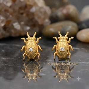 May include: A pair of gold-toned beetle-shaped earrings, each featuring a textured surface and a single small clear round gemstone set in the center of the back. The text GemstonetownDesign is visible in the lower right corner of the image.