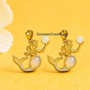May include: Two gold-toned mermaid pendants featuring a central oval iridescent opal stone on the tail and a smaller round white opal sphere held in the hand. The mermaid tail is accented with small clear stones. Text reads GemstoneTownDesign.