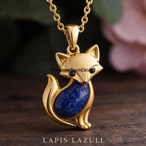 Puede incluir: Un colgante de zorro en tono dorado con una incrustación de piedra lapislázuli azul profundo en el cuerpo y pequeños ojos de cuentas negras. El zorro está representado en posición sentada, suspendido de una cadena de eslabones dorada.