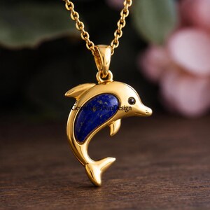 Op de afbeelding: Een goudkleurige dolfijn-hanger met een diepblauwe lapis lazuli inleg op het lichaam en een klein zwart oogdetail. De hanger hangt aan een bijpassende goudkleurige ketting en bevat de tekst GemstoneTownDesign op het oppervlak.