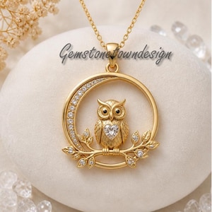 Puede incluir: Collar con colgante de búho dorado. El búho está posado en una rama dentro de un marco circular, adornado con cristales transparentes. El búho tiene un cristal en forma de corazón en el pecho. El collar tiene el texto "Gemstonetowndesign".