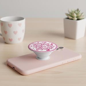 May include: A white PopSocket with a pink and white design that says "Come On Barbie Let's Go Party!" The PopSocket is attached to a pink smartphone. A white mug with pink hearts is in the background.