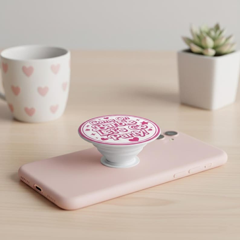 May include: A white PopSocket with a pink and white design that says "Come On Barbie Let's Go Party!" The PopSocket is attached to a pink smartphone. A white mug with pink hearts is in the background.