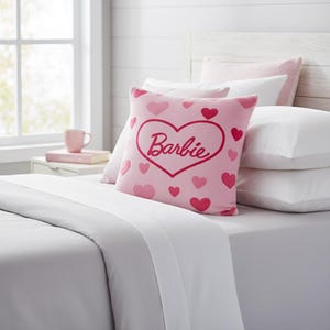 May include: A pink decorative pillow with a heart-shaped design and the name "Barbie" in cursive. The pillow is adorned with various pink hearts. The pillow is placed on a bed with white sheets and pillows, creating a soft and inviting aesthetic.
