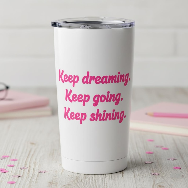 May include: White travel tumbler with a black lid. The tumbler features the pink text "Keep dreaming. Keep going. Keep shining." The tumbler is on a light-colored wooden surface with pink confetti.