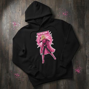 May include: Black hooded sweatshirt featuring a pink and black graphic of Super Barbie. The graphic includes the text "SUPER BARBIE" in a white rectangular box. The design shows Barbie in a superhero pose with a pink cape.