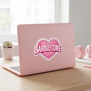 May include: A pink laptop with a sticker featuring the word "BARBIECORE" inside a heart shape. The heart has a textured pink background with two yellow lightning bolts. The laptop is on a wooden desk.