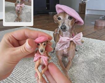 May include: A small Italian Greyhound dog wearing a pink beret and a matching pink satin bow collar, positioned next to a miniature figurine of the same dog wearing an identical pink beret and bow, held in a person's hands.