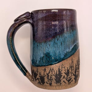 May include: A handmade ceramic mug with a unique design. The top half is glazed in shades of purple and teal, while the bottom features a textured, unglazed finish with a black tree line pattern. The handle is a dark teal color.