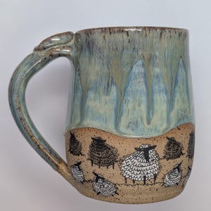 May include: A ceramic mug featuring a light blue and tan glaze with a drip pattern. The lower portion displays a hand-drawn illustration of multiple sheep in black and white ink against a speckled tan background with a curved handle.