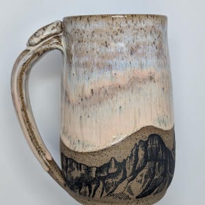 May include: A handmade ceramic mug with a mountain design. The mug features a light brown base with a black mountain range illustration. The upper portion has a gradient of pink, white, and brown. The handle is a light brown color.