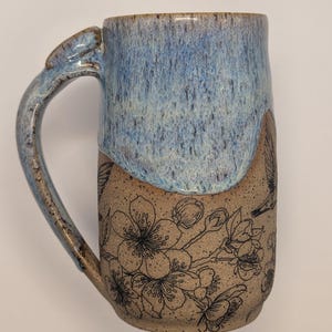 May include: A handmade ceramic mug with a blue glazed top and a tan base. The base features a black floral design. The mug has a large handle and is suitable for hot or cold beverages.