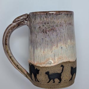 May include: A ceramic mug featuring a speckled tan base with black silhouettes of cats walking around the bottom. The upper portion of the mug has a smooth, dripped glaze effect in shades of mauve, lavender, and cream with a glossy finish.