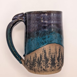 May include: A ceramic mug featuring a two-tone glaze design. The upper portion is finished in a glossy deep purple and teal drip glaze, while the lower portion displays a natural speckled tan clay body with a black printed forest tree pattern.