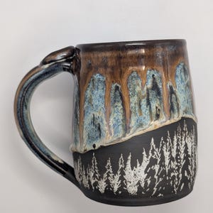 May include: A ceramic mug featuring a dark matte black base with a white etched forest of pine trees. The upper portion of the mug has a glossy glaze that transitions from a light blue to a deep brown, creating a drip effect over the black base.