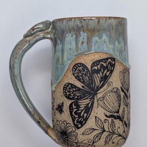 May include: A ceramic mug with a speckled tan base and a blue drip glaze at the top. The exterior features black line art illustrations of a large butterfly, various flowers, and a small bee. The mug includes a curved handle with a matching blue glaze.