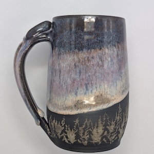 May include: A large ceramic mug with a dark blue and purple glaze. The top half of the mug has a gradient effect, transitioning to a black base with a forest silhouette design. The handle is also glazed.
