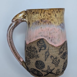 May include: A ceramic mug with a speckled tan and brown base featuring black line art illustrations of seashells, starfish, and coral. The upper portion of the mug has a smooth, glossy glaze transition from light pink to a speckled tan and brown.