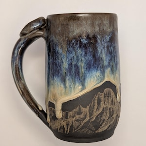 May include: A handmade ceramic mug with a mountain design. The mug features a dark brown top that fades into blue and cream colors. The handle has a unique, textured design. The base of the mug is black with a mountain range etched into the surface.