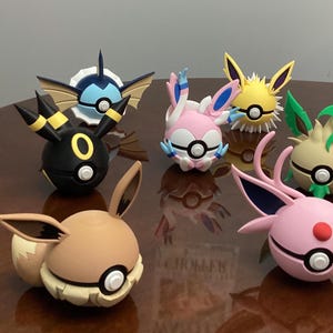May include: A collection of colorful, stylized figures resembling Pokémon characters. The figures are in various colors, including orange, black, pink, yellow, green, and blue, and are arranged on a wooden surface.
