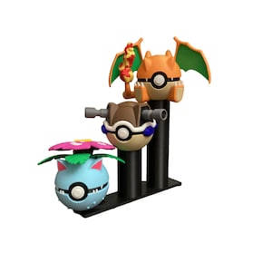 May include: A display stand featuring three colorful Pokémon-themed figures. The figures include a blue and green Venusaur, a brown and white figure, and an orange Charizard with green wings. The figures are mounted on black cylindrical supports.