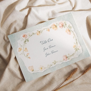 Pode incluir: Um cartão de mesa com uma borda floral. O cartão diz "Table One, Jane Brown, John Brown". O cartão tem um fundo azul claro e uma borda floral decorativa. Sobre uma superfície de linho bege.