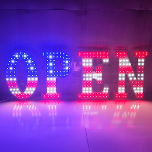 May include: An illuminated sign spelling "OPEN" in large, block letters. The "O" is blue with white dots, and the "PEN" is red and white, resembling the American flag. The sign is lit with LED lights.