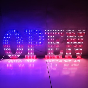 May include: An illuminated sign spelling "OPEN" in large, block letters. The letters are composed of numerous small, bright LED lights, with a gradient of colors from blue to red. The sign is likely used for business advertising.