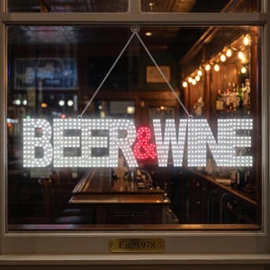 May include: A rectangular LED sign hanging by silver chains in a window, displaying the text BEER & WINE in bright white light, with the ampersand symbol illuminated in red. The sign is composed of individual light dots forming the letters.