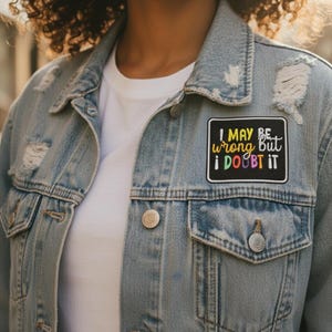 May include: Light blue denim jacket with distressed details and a white t-shirt underneath. A black patch on the jacket reads "I MAY BE wrong but I DOUBT IT" in colorful text. The jacket has silver buttons and two front pockets.