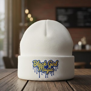 May include: A white knit beanie with the embroidered text "Black Girl Magic" in yellow and blue. The text has a dripping effect. The beanie is sitting on a wooden surface.