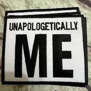 Puede incluir: Parche rectangular blanco con borde negro y texto "UNAPOLOGETICALLY ME" en letras negras en negrita. El parche es de tela y está diseñado para ser adherido a ropa o accesorios.