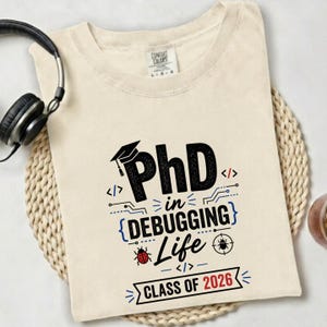 May include: Cream-colored t-shirt with the text "PhD in Debugging Life Class of 2026" in black and blue. The design includes a graduation cap, code symbols, and a ladybug and a bug icon. A pair of black headphones rests on the side.