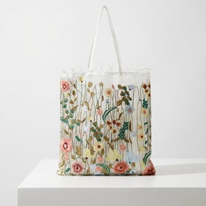 Puede incluir: Un bolso tote blanco con un diseño de bordado floral. El bolso presenta una variedad de flores coloridas en tonos de rosa, amarillo, azul y verde. El bolso tiene dos asas blancas y un ribete decorativo en la parte superior.