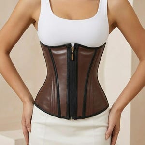 May include: A brown corset with a black zipper and trim. The corset is made of a smooth material and has a structured design. The zipper is centered and runs the length of the corset. The corset is worn over a white tank top and paired with a white skirt.