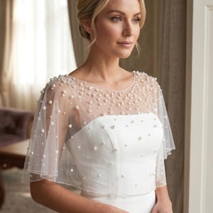 K&ouml;nnte beinhalten: Ein wei&szlig;es Braut-Capelet mit einem transparenten T&uuml;ll&uuml;berwurf, der mit verstreuten wei&szlig;en Perlen unterschiedlicher Gr&ouml;&szlig;e verziert ist. Das Capelet hat einen runden Ausschnitt und kurze, flie&szlig;ende &Auml;rmel, die &uuml;ber einem schlichten wei&szlig;en Kleid liegen.