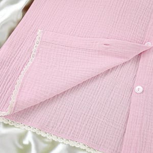 Puede incluir: Una camisa abotonada de color rosa claro hecha de una tela texturizada y arrugada. La prenda presenta un delicado borde de encaje blanco a lo largo de la tapeta frontal y el dobladillo, con peque&ntilde;os botones blancos a lo largo de la abertura.