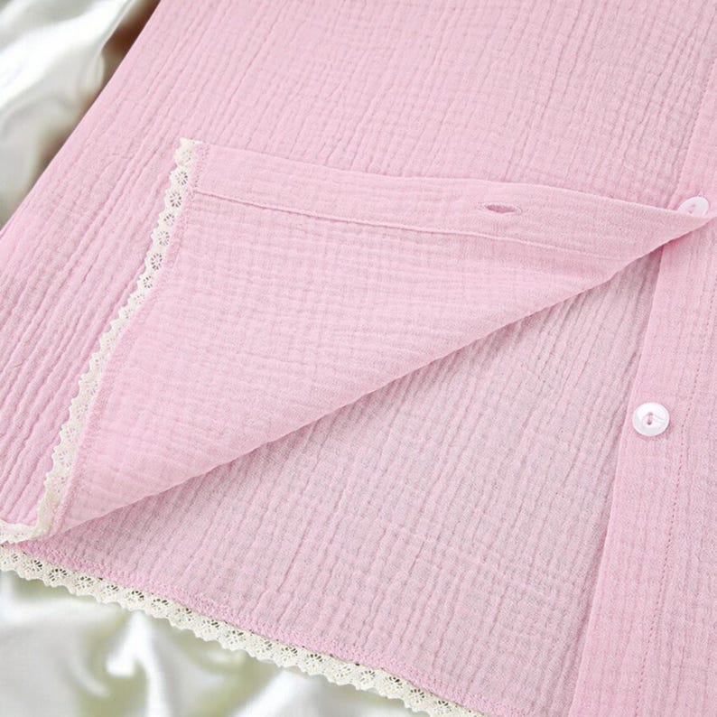Puede incluir: Una camisa abotonada de color rosa claro hecha de una tela texturizada y arrugada. La prenda presenta un delicado borde de encaje blanco a lo largo de la tapeta frontal y el dobladillo, con peque&ntilde;os botones blancos a lo largo de la abertura.