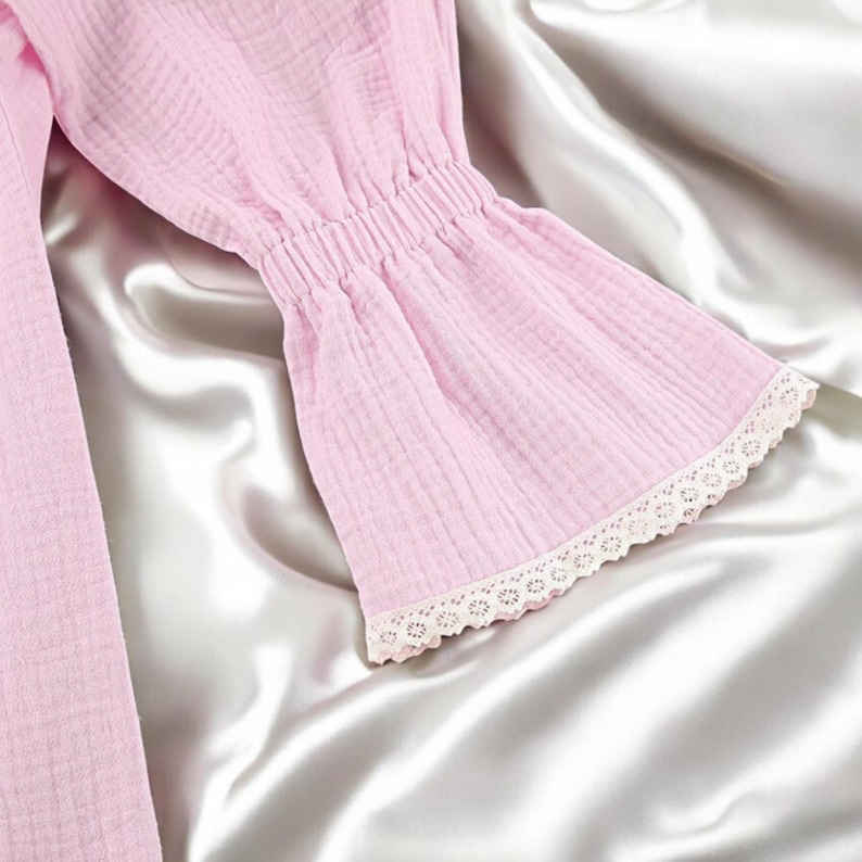 Puede incluir: Una prenda de manga larga en color rosa claro hecha de tela texturizada y arrugada. La manga presenta un pu&ntilde;o fruncido y el&aacute;stico que se ensancha en forma de campana, rematado con un delicado borde de encaje blanco en el extremo.