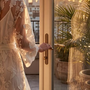 K&ouml;nnte beinhalten: Ein wei&szlig;es Spitzenkleid mit passendem G&uuml;rtel und Unterkleid. Der Morgenmantel hat lange, ausgestellte &Auml;rmel und wird ge&ouml;ffnet gehalten. Der Hintergrund zeigt eine Skyline und Topfpflanzen.