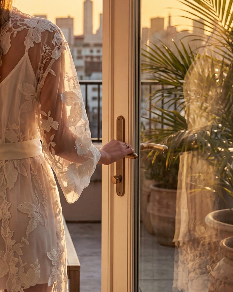K&ouml;nnte beinhalten: Ein wei&szlig;es Spitzenkleid mit passendem G&uuml;rtel und Unterkleid. Der Morgenmantel hat lange, ausgestellte &Auml;rmel und wird ge&ouml;ffnet gehalten. Der Hintergrund zeigt eine Skyline und Topfpflanzen.