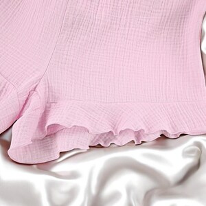 Puede incluir: Unos pantalones cortos de color rosa claro confeccionados con un tejido texturizado y arrugado. La prenda presenta un dobladillo con volantes a lo largo del borde inferior, creando un aspecto suave y superpuesto con un patr&oacute;n de tejido visible.