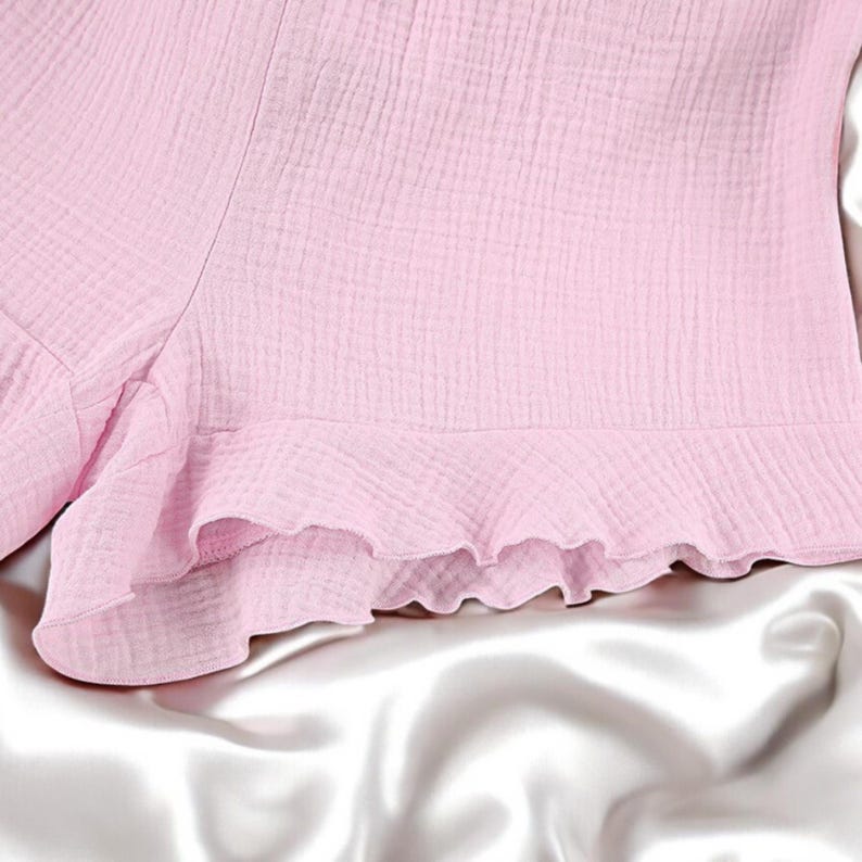 Puede incluir: Unos pantalones cortos de color rosa claro confeccionados con un tejido texturizado y arrugado. La prenda presenta un dobladillo con volantes a lo largo del borde inferior, creando un aspecto suave y superpuesto con un patr&oacute;n de tejido visible.