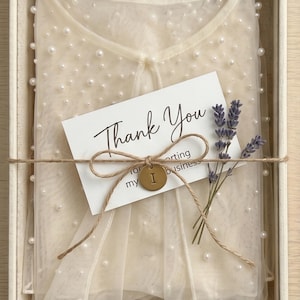 May include: A cream-coloured sheer mesh top embellished with scattered white pearls, folded inside a box. A white card with the text Thank You is tied to the top with twine, featuring a small brass-coloured circular tag with the letter I and a sprig of dried lavender.