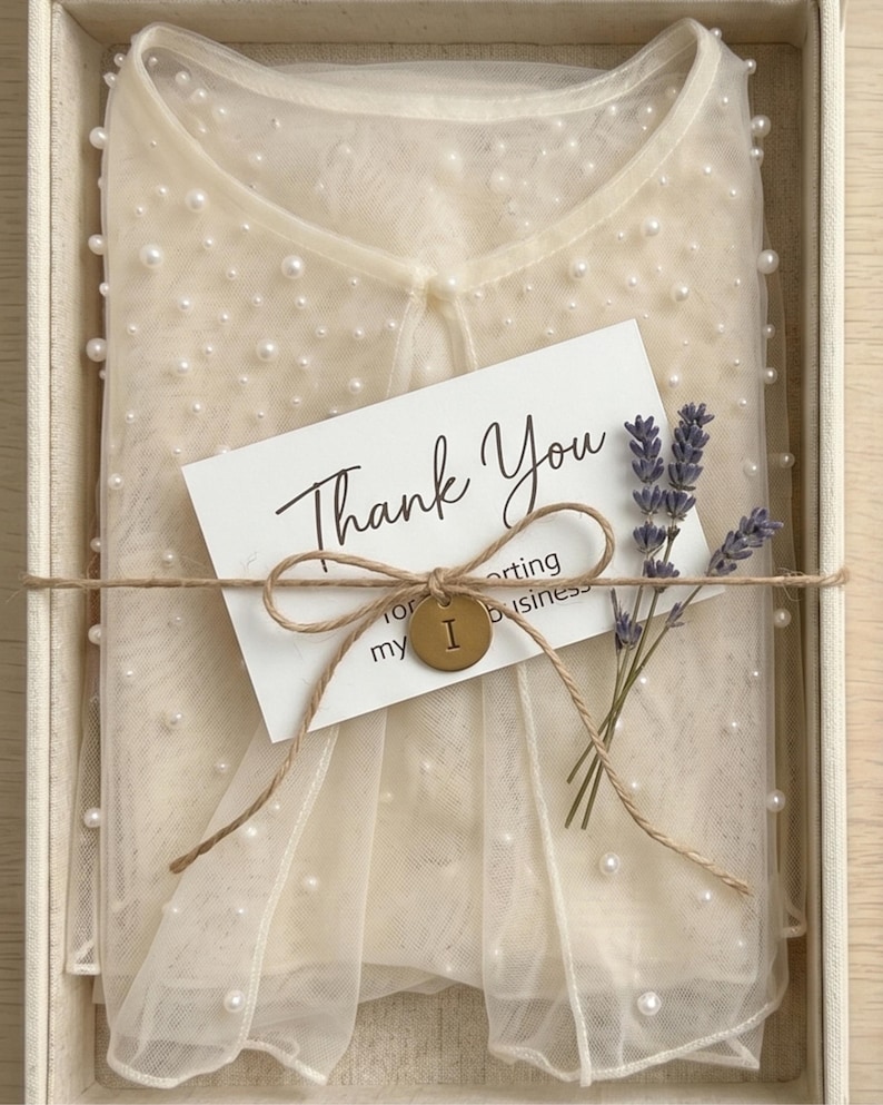 May include: A cream-coloured sheer mesh top embellished with scattered white pearls, folded inside a box. A white card with the text Thank You is tied to the top with twine, featuring a small brass-coloured circular tag with the letter I and a sprig of dried lavender.