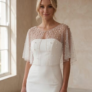 K&ouml;nnte beinhalten: Ein transparentes wei&szlig;es T&uuml;ll-Capelet mit verstreuten wei&szlig;en Perlen in verschiedenen Gr&ouml;&szlig;en auf dem Stoff. Das Capelet hat einen runden Ausschnitt, ein offenes Vorderteil und einen zarten, perlenbesetzten Saum entlang der Kanten.