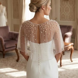K&ouml;nnte beinhalten: Ein bodenlanges wei&szlig;es Kleid mit einem transparenten T&uuml;ll-Capelet, das mit verstreuten wei&szlig;en Perlen besetzt ist. Das Capelet hat ein offenes R&uuml;ckendesign und das Kleid verf&uuml;gt &uuml;ber eine vertikale Reihe kleiner wei&szlig;er Kn&ouml;pfe am Rei&szlig;verschluss.