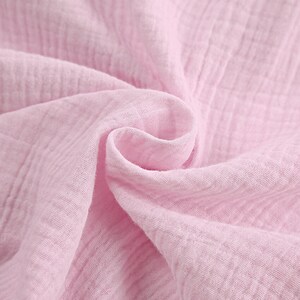 Puede incluir: Una tela de color rosa claro suave con una textura arrugada caracter&iacute;stica de la doble gasa. El material est&aacute; drapeado en pliegues sueltos y fluidos, resaltando el patr&oacute;n de cuadr&iacute;cula del tejido y su color pastel s&oacute;lido y uniforme.