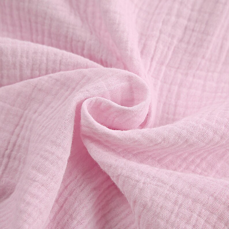Puede incluir: Una tela de color rosa claro suave con una textura arrugada caracter&iacute;stica de la doble gasa. El material est&aacute; drapeado en pliegues sueltos y fluidos, resaltando el patr&oacute;n de cuadr&iacute;cula del tejido y su color pastel s&oacute;lido y uniforme.